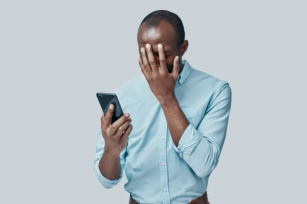 Sfrustrowany Młody Afrykanin Pomocą Smartfona Zakrywając Twarz Ręką Stojąc Szarym — Zdjęcie stockowe