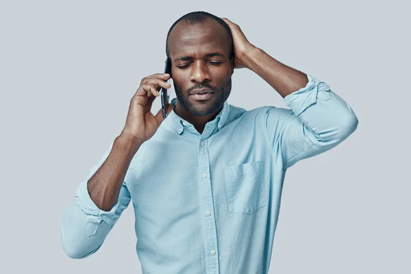 Frustrated young African man talking on the smartphone while standing against grey background