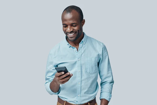 年轻英俊的非洲男人 站在灰色的背景下 一边用智能手机一边笑 — 图库照片