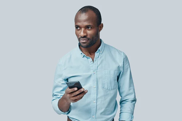 Przystojny Młody Afrykanin Pomocą Smartfona Stojąc Szarym Tle — Zdjęcie stockowe