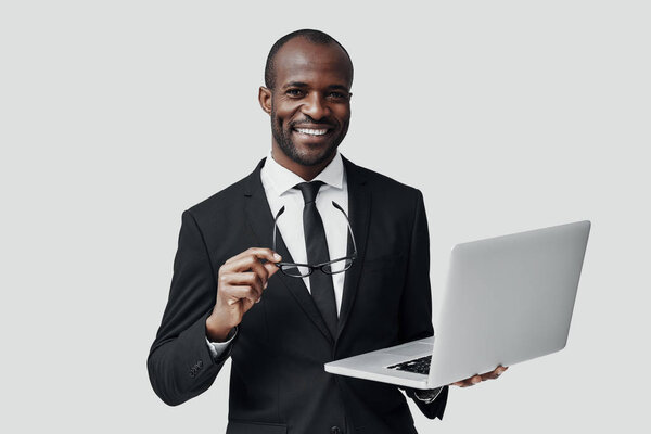 Happy young African man in formal wear working using computer while standing against grey background     