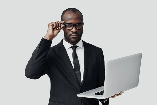 穿着正装的严肃的非洲年轻人站在灰色背景下 一边用电脑干活 — 图库照片