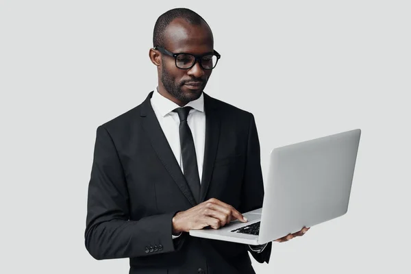 身穿正装的忙碌的非洲年轻人站在灰色背景下 一边用电脑干活 — 图库照片