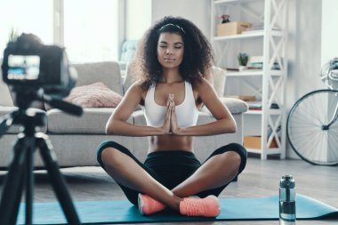 Sosyal medya videosu çekerken yoga pozisyonunda oturan güzel Afrikalı genç bir kadın.             