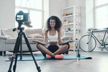 Çekici genç Afrikalı kadın yoga pozisyonunda oturuyor ve sosyal medya videosu çekerken gülümsüyor.    
