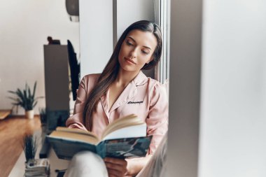 Evdeki pencere pervazında dinlenirken rahat pijamalarıyla kitap okuyan güzel genç bir kadın.                  