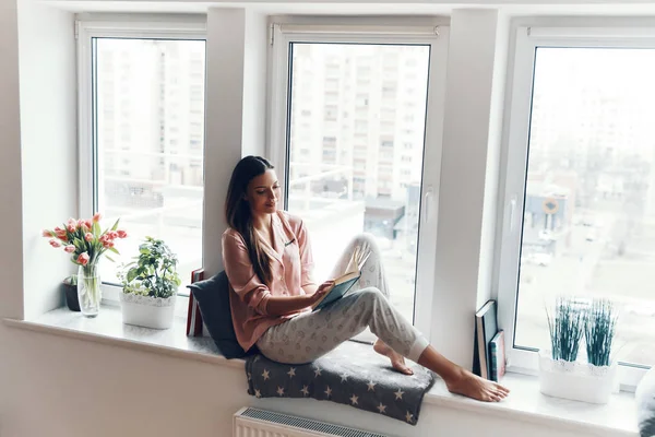 Vonzó Fiatal Kényelmes Pizsamában Könyvet Olvas Miközben Otthon Pihen Ablakpárkányon — Stock Fotó