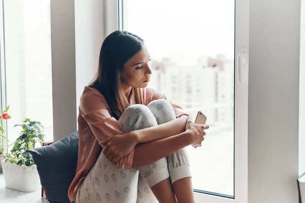 穿着舒适睡衣 头脑清醒的年轻女人 一边拿着智能手机 一边在家里的窗台上休息 — 图库照片