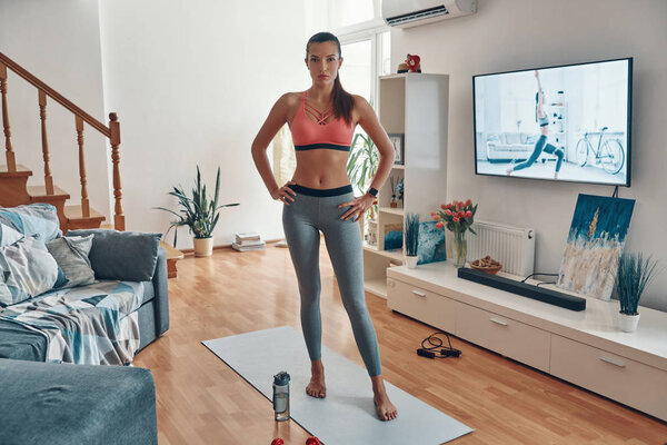 Full length of beautiful fit young woman in sports clothing looking at camera while working out in the living room 