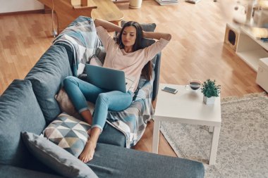 Gündelik kıyafetlerle evde vakit geçirirken dizüstü bilgisayar kullanan çekici genç bir kadın.