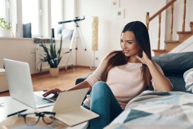 Dizüstü bilgisayar kullanan ve evde dinlenirken gülümseyen sıradan giysili hoş bir genç kadın.        