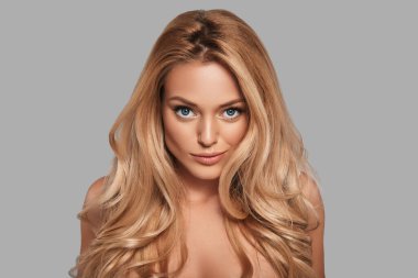 Taze parlak tenli şehvetli genç bir kadın kameraya bakıyor ve gri arka planda dururken gülümsüyor.