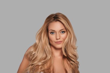 Taze parlak ciltli çekici genç bir kadın kameraya bakıyor ve gri arka planda dururken gülümsüyor.