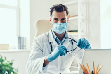 Beyaz laboratuvar önlüklü ve koruyucu maskeli kendinden emin genç adam içeride otururken kameraya bakıyor.