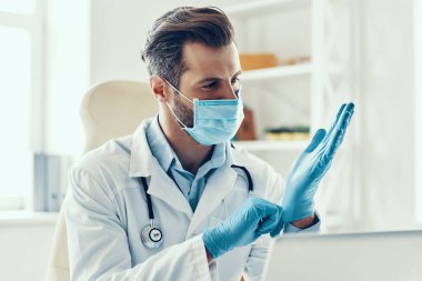 Beyaz laboratuvar önlüklü ve koruyucu maskeli genç adam içeride otururken eldiveni ayarlıyor.