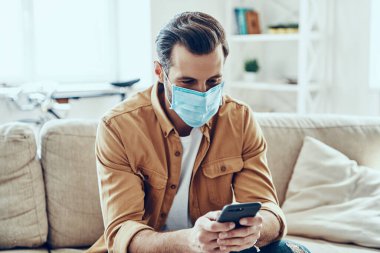 Evde çalışırken akıllı telefon kullanan koruyucu maskeli genç adam.