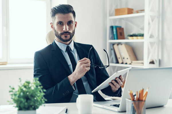 Good looking young man in elegant business suit working using digital tablet and looking at camera while sitting in the office