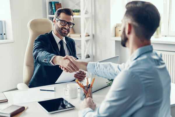 Two happy young men in formal wear shaking hands and smiling while sitting in the office