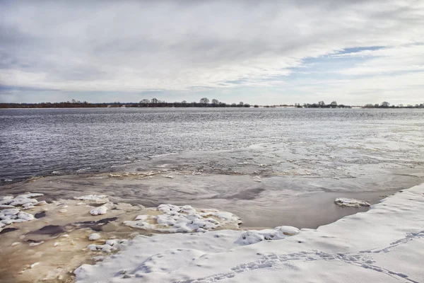 Kış nehir kaplı kar ve buz kıyıya, bulutlu bir gökyüzü ve ufukta ağaçlar altında tarafından