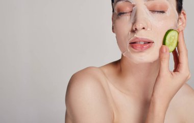 Nazik, sofistike, nemlendirici maskeli, arka planda taze bir salatalık olan sakin bir kızı kapat. Gözü kapalı ve sol eli salatalığa dokunuyor.