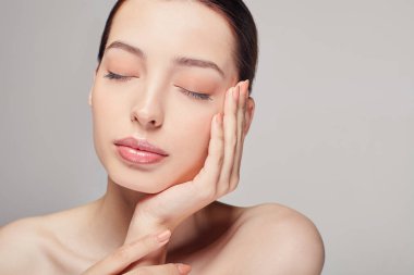 Kahverengi saçlı, temiz tenli, çıplak omuzlu güzel bir kız yüzünü gri stüdyo arka planında poz vermiş, kapalı gözlü, açık renk makyajlı bir model.