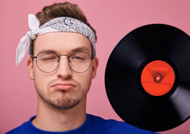 Beyaz bandanalı, koyu mavi kazaklı, pembe arka planda duran havalı adam. 