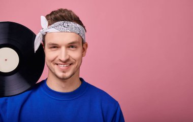 Beyaz bandanalı, koyu mavi kazaklı, pembe arka planda duran havalı adam. 