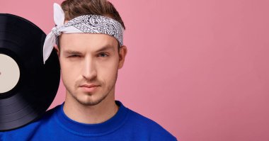 Beyaz bandanalı, koyu mavi kazaklı, pembe arka planda duran havalı adam. 
