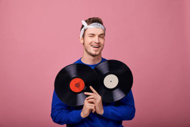 Beyaz bandanalı, lacivert kazaklı, havalı bir adam. Pembe arka planda plaklar var. 