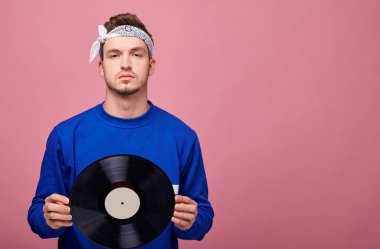 Beyaz bandanalı, koyu mavi kazaklı, pembe arka planda duran havalı adam. 