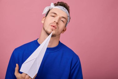 Koyu mavi kazaklı adam elinde kağıt uçakla pembe arka planda duruyor.