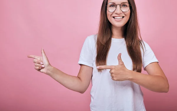 Joyful girl in T-shirt and round glasses shows forefingers to side — Stock Photo, Image