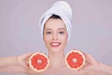 A smiling girl in a white towel on her head holds a cut grapefruit near her face.