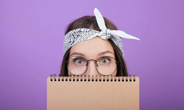En söt brunhårig kvinna i runda glasögon döljer sitt ansikte bakom en anteckningsbok. — Stockfoto