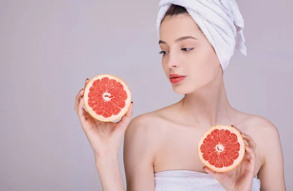 Young attractive girl with clean skin, half-naked, in a towel on her head, holds in her hands a fresh cut grapefruit. — Stock Photo, Image