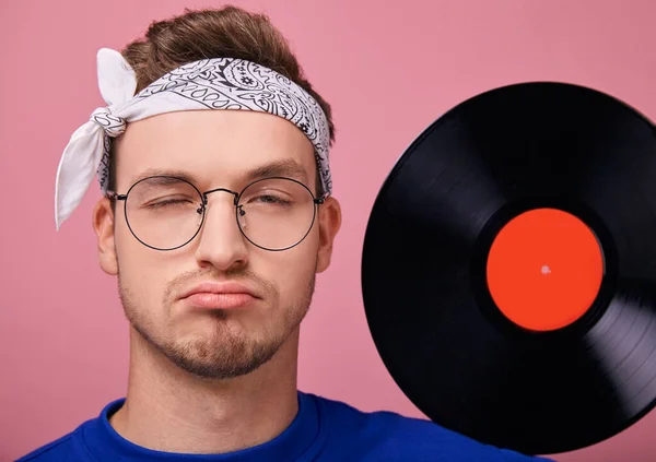 Alegre chico hippie humorístico con los labios doblados en gafas con sostiene disco de vinilo negro con una etiqueta roja — Foto de Stock