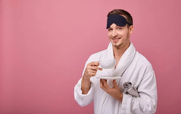 Sleepy man in a sleeping mask on head in a white coat with a newspaper under his arm, smiling at the camera with a cup of coffee in hands. Awakening. Copy space. Looking straight with smile.