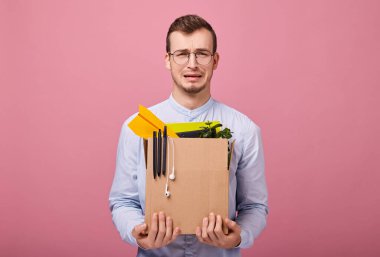 Kutuya odaklan. Gökyüzü mavisi gömlekli ve bilgisayar gözlüklü genç bir adam pembe arka planda elinde kalem, bitki ve kulaklık olan bir karton kutunun üzerinde duruyor. Kovuldun. Adam üzgün..