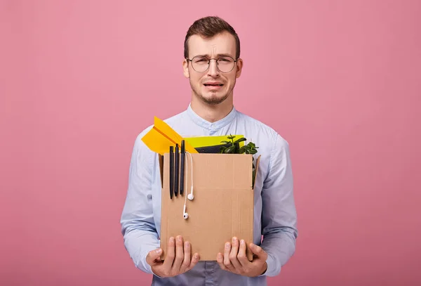 Focus at the box. A young pretty guy in a sky blue shirt and computer glasses stands on a pink background with a cardboard box with pens, plant and headphones in his hands. Fired. Man is upset.