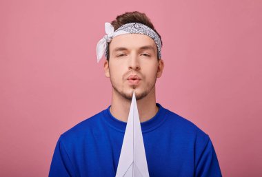 Kapatın. Koyu mavi kazaklı bandana giyen beyaz giysili sakin adam elinde beyaz kağıt uçakla duruyor, bu da yan tarafı gösteriyor. Uçakta üfleyen bir çocuk. Boşluğu kopyala.