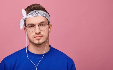 Gözlüklü, beyaz bandanalı, mavi ceketli, müzik dinleyen sakin sakin bakan kendine güvenen adam. Pembe arkaplan, kopyalama alanı. 