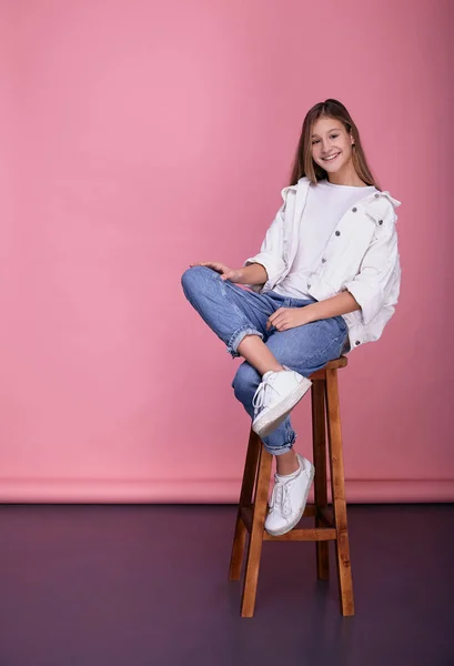 Chica joven vestida a la moda se sienta en una silla sobre un fondo rosa . —  Fotos de Stock