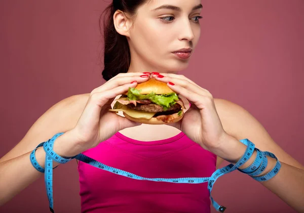 Beautiful girl with a measuring tape holds a burger. — Stock Photo, Image