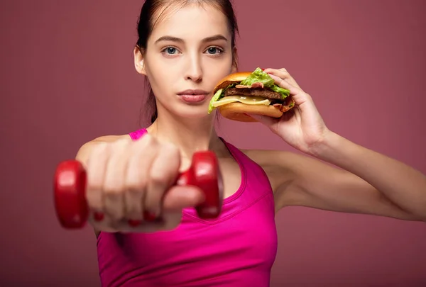 En slank dame spiser et smørbrød og holder dumklokker . – stockfoto