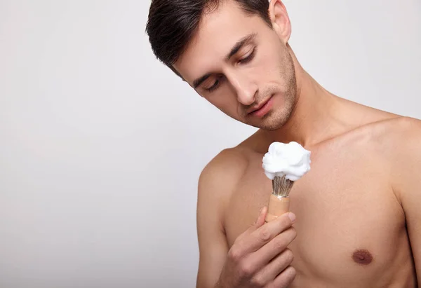 Ein gutaussehender Mann mit nacktem Oberkörper wird Rasierschaum auf sein Gesicht auftragen. — Stockfoto