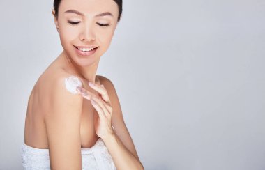 A young Asian woman puts body cream on her bare shoulder.