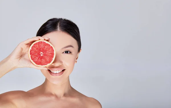 Seorang wanita Asia muda yang tersenyum memegang jeruk di dekat wajahnya . — Stok Foto