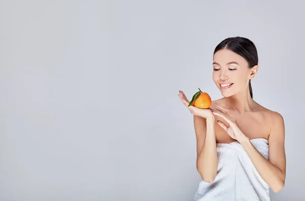 Fresh cute Asian model in a towel with orange in hand on a grey background. — Stock Photo, Image