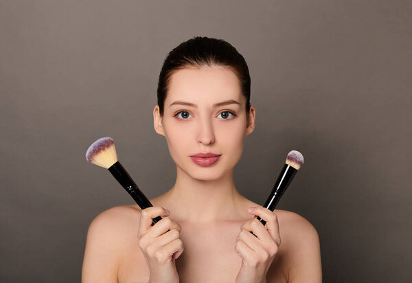 A young beautiful white girl with bare shoulders holds makeup brushes.