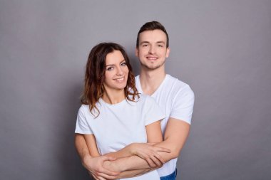 İki genç, yakışıklı, çekici, neşeli, pozitif çiftin portresi, insanlar gülüşüyor, beyaz temel tişörtler giyiyor, gri arka planda izole edilmiş. Boşluğu kopyala.